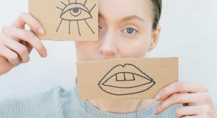 Frau hält vor ihrem Auge und vor dem Mund mit einem Auge und einem Mund bemalte Pappstückchen.