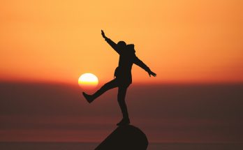 Mann balanciert auf einem Felsen mit ausgestrecktem Bein vor untergehender Sonne, es sieht aus, als ob die Sonne, wie einen Ball, mit dem Fuß trifft.