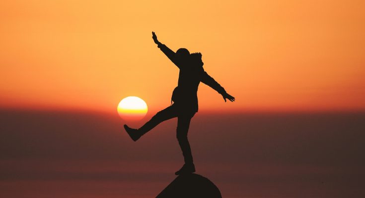 Mann balanciert auf einem Felsen mit ausgestrecktem Bein vor untergehender Sonne, es sieht aus, als ob die Sonne, wie einen Ball, mit dem Fuß trifft.