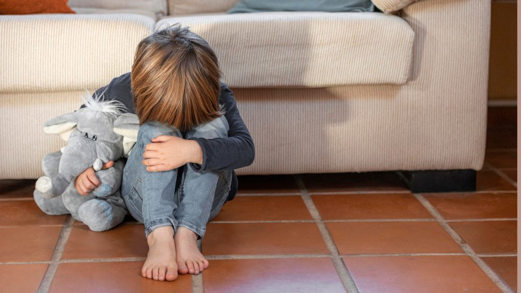 Ein Junge sitzt neben dem Sofa auf dem Boden, den Kopf auf die Knie gelegt.