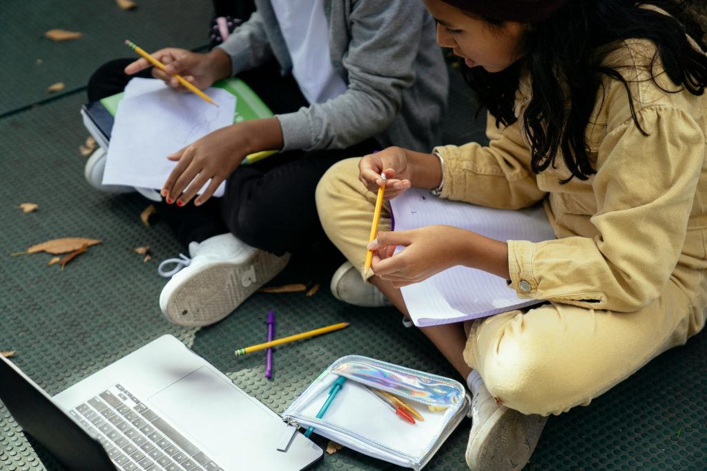 Zwei Schulkinder sitzen im Schneidersitz nebeneinander mit Stiften und Heften.