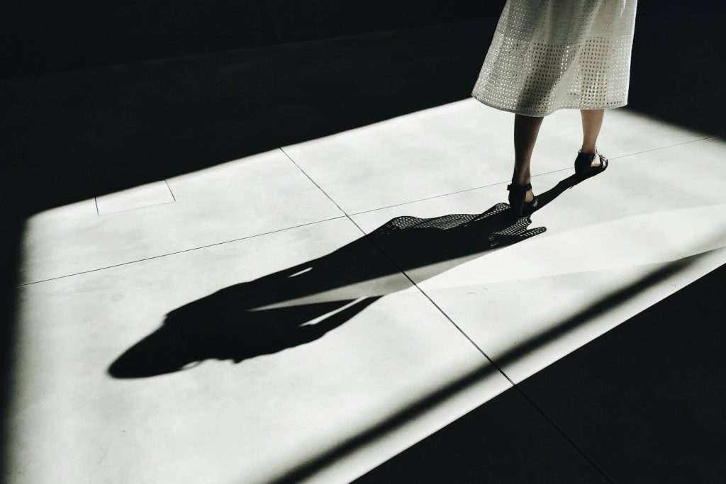 Frauenbeine in einem weißen Rock gehen aus dem Bild hinaus, dabei werfen die Beine einen Schatten.