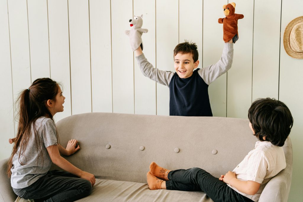 Ein Junge führt mit zwei Handpuppen ein Kindertheater vor, zwei andere Kinder schauen ihn lachend an. 